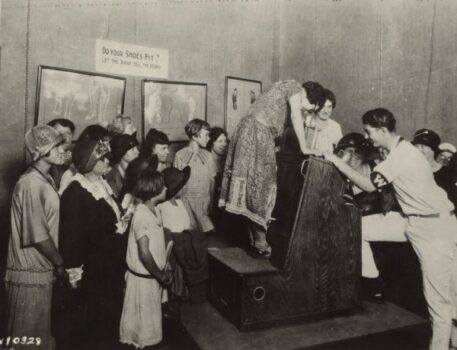 Elképesztő történelem: nők és gyerekek csoportja vesz körbe egy nőt, aki egy fluoroszkópon állva veszi szemügyre, megfelelően állnak-e a lábai a felhúzott cipőben (1926)